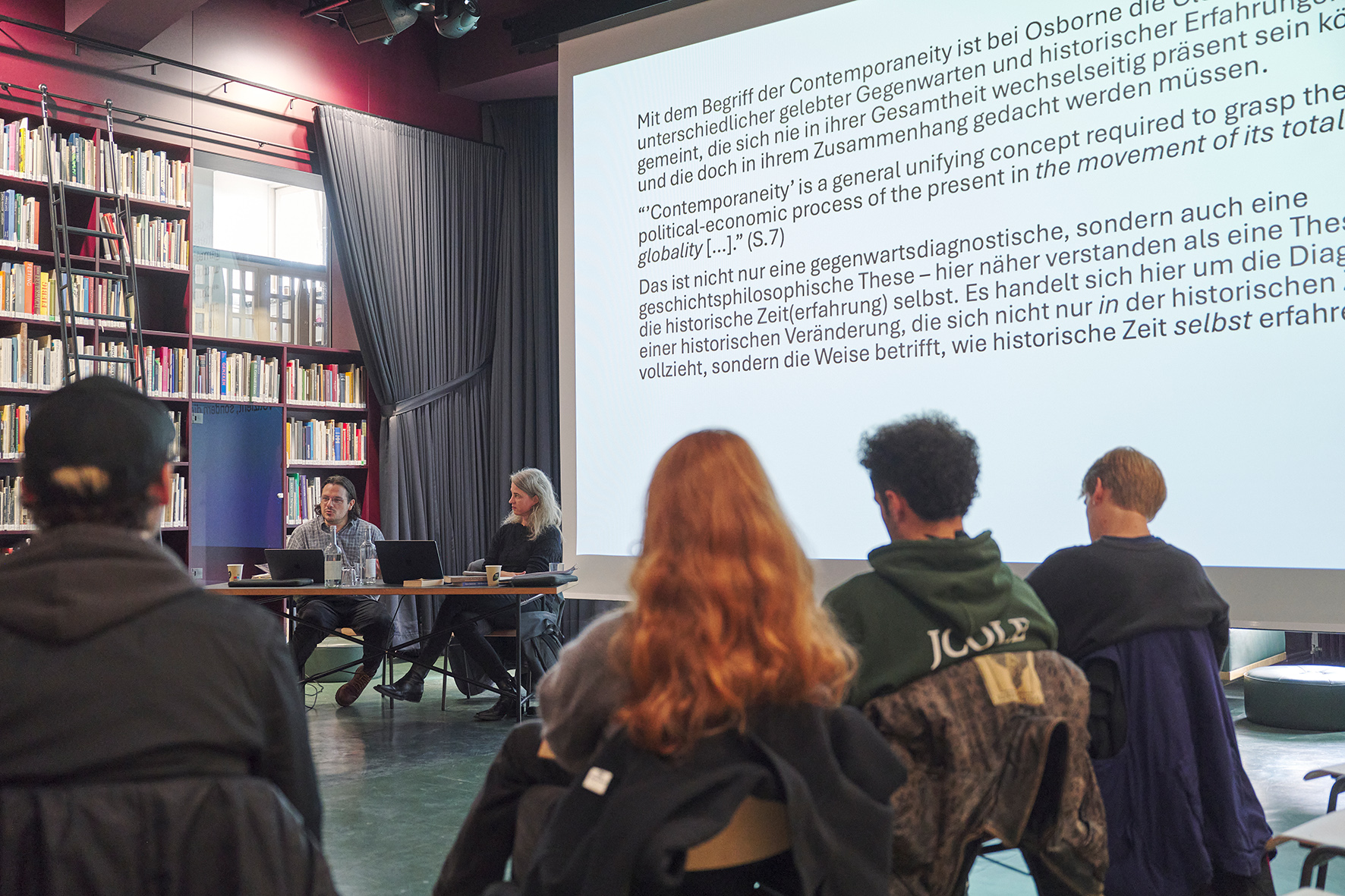 Vortragssituation in einer Bibliothek: Zwei Personen sitzen an einem Tisch mit Laptops und sprechen vor einem Publikum. Hinter ihnen wird eine Präsentationsfolie mit einem längeren Text auf Deutsch und Englisch projiziert. Bücherregale und hohe Fenster rahmen den Raum, das Publikum ist im Vordergrund von hinten zu sehen.