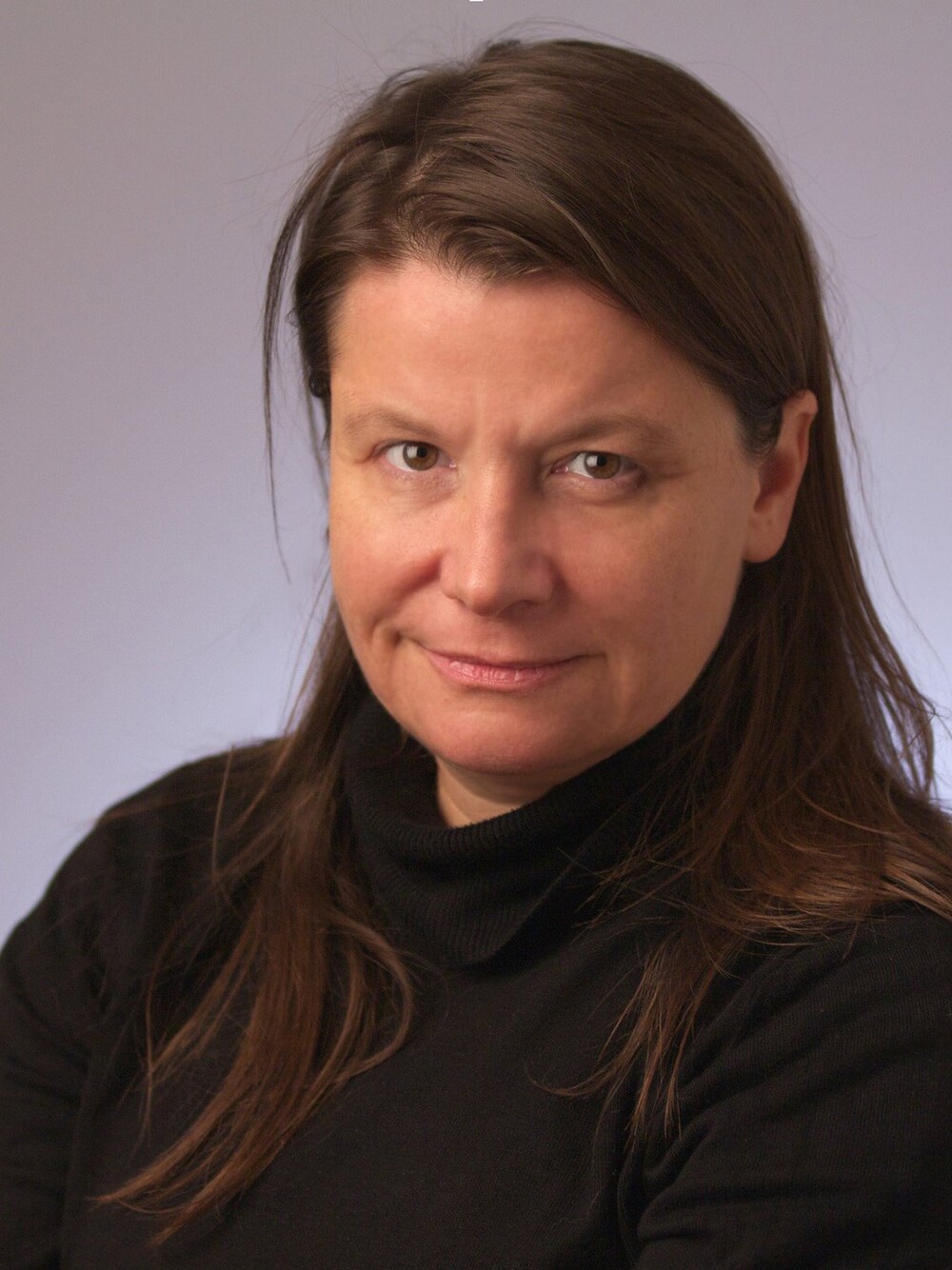 Portrait of a woman with long hair wearing a black turtleneck, photographed against a neutral gray background.