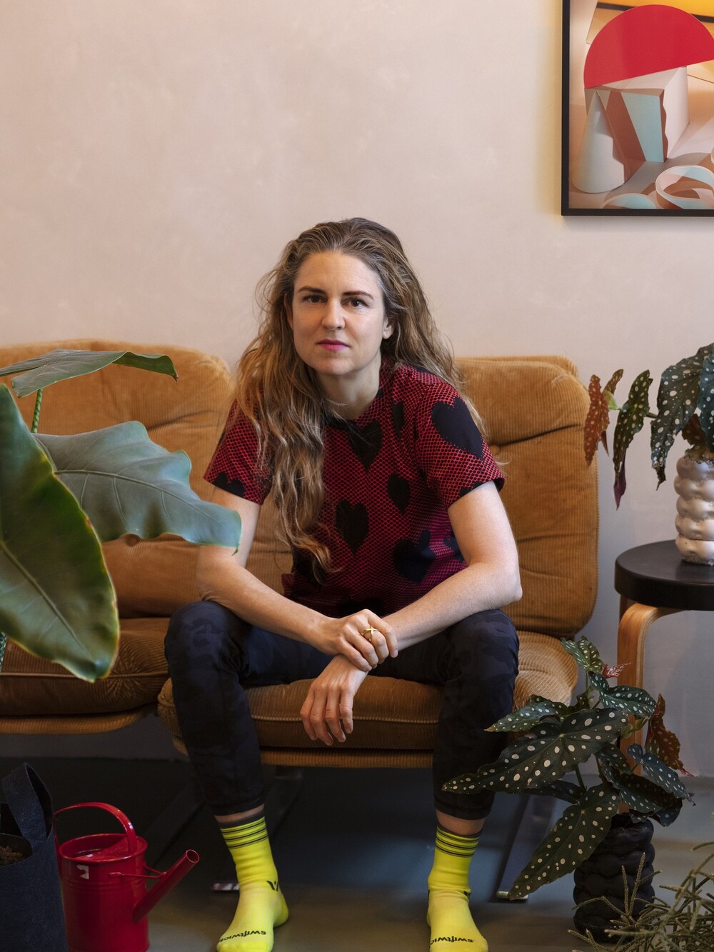 Eine Frau mit langen blonden gewellten Haaren sitzt mit leicht geöffneten Beinen auf einem Sofa, sie stützt ihre Arme gebaugt auf ihren Knien ab. Sie trägt neongelbe Socken, eine blaue Jeans und ein rotes Shirt mit schwarzen großen Herzen. Topfpflanzen rahmen sie auf dem Sofa ein.