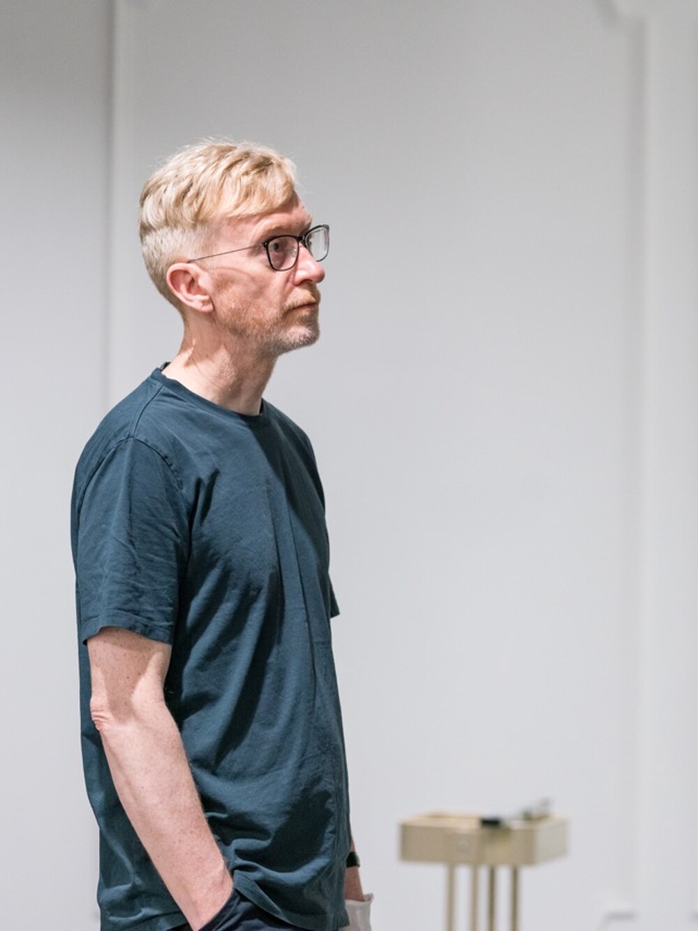 Mann mit kurzen blonden Haaren und Brille steht im Profil in einem Ausstellungsraum mit weißen Wänden.