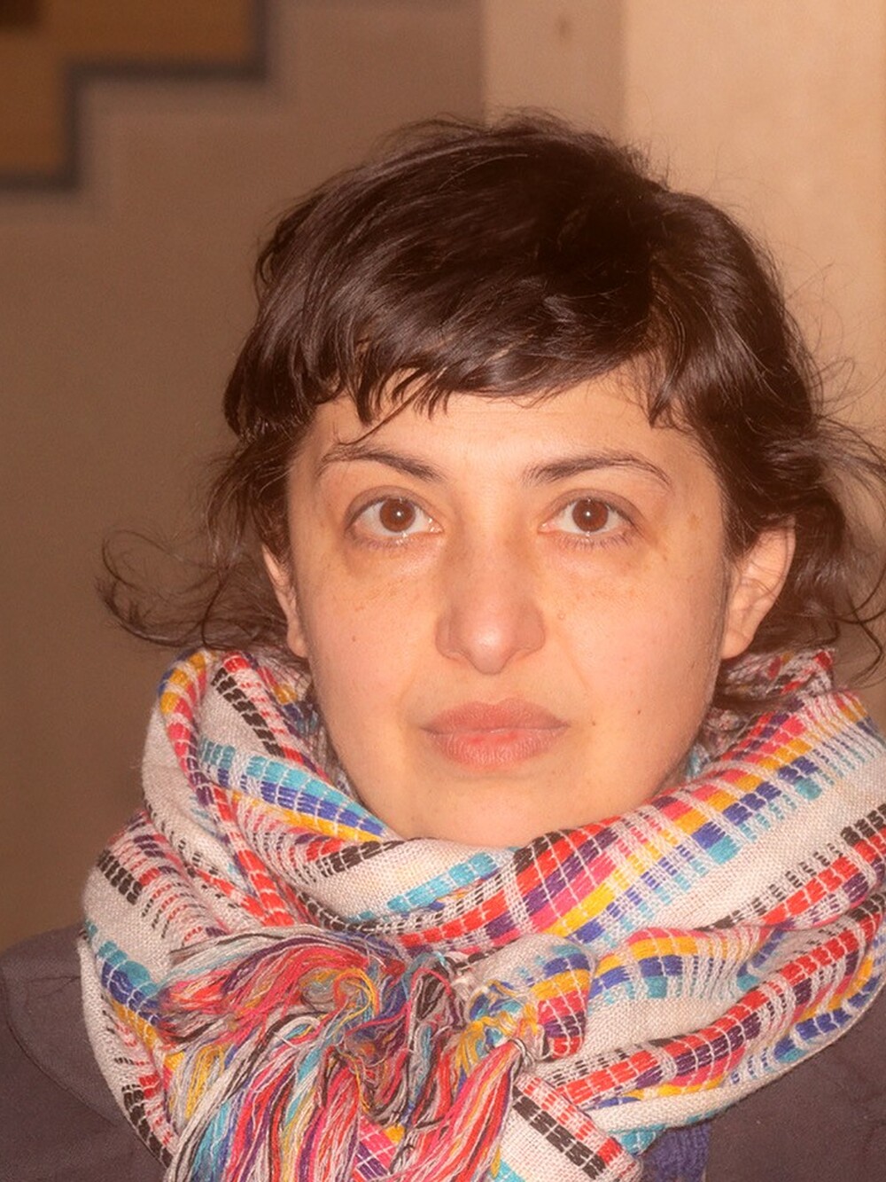 Portrait of a person with short dark hair looking directly at the camera. They are wearing a thick, multicolored patterned scarf and standing indoors with a staircase in the background.