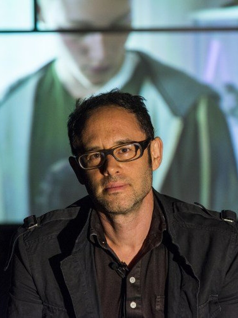A middle-aged man wearing glasses and a dark jacket is looking into the camera. He has short black hair.