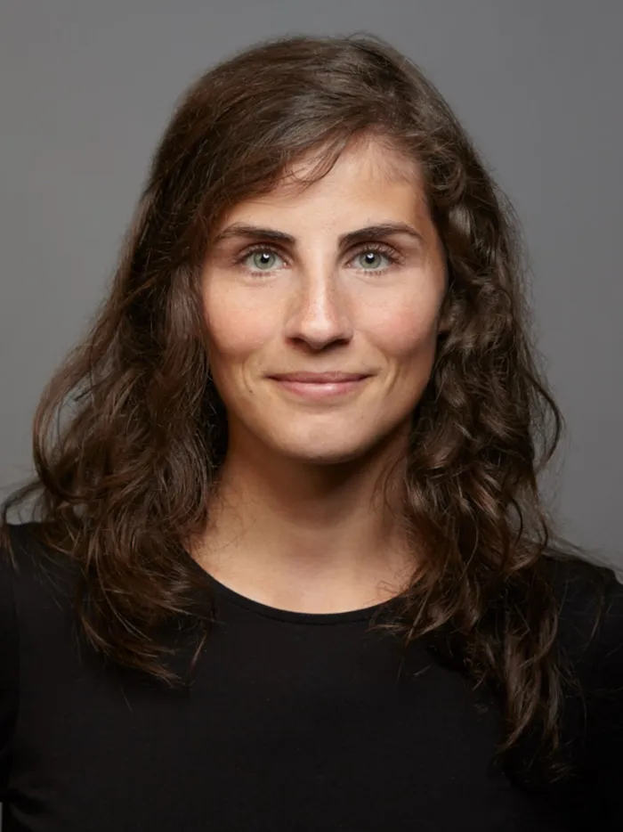 Portrait of a woman with curly hair and a neutral expression, against a grey background.