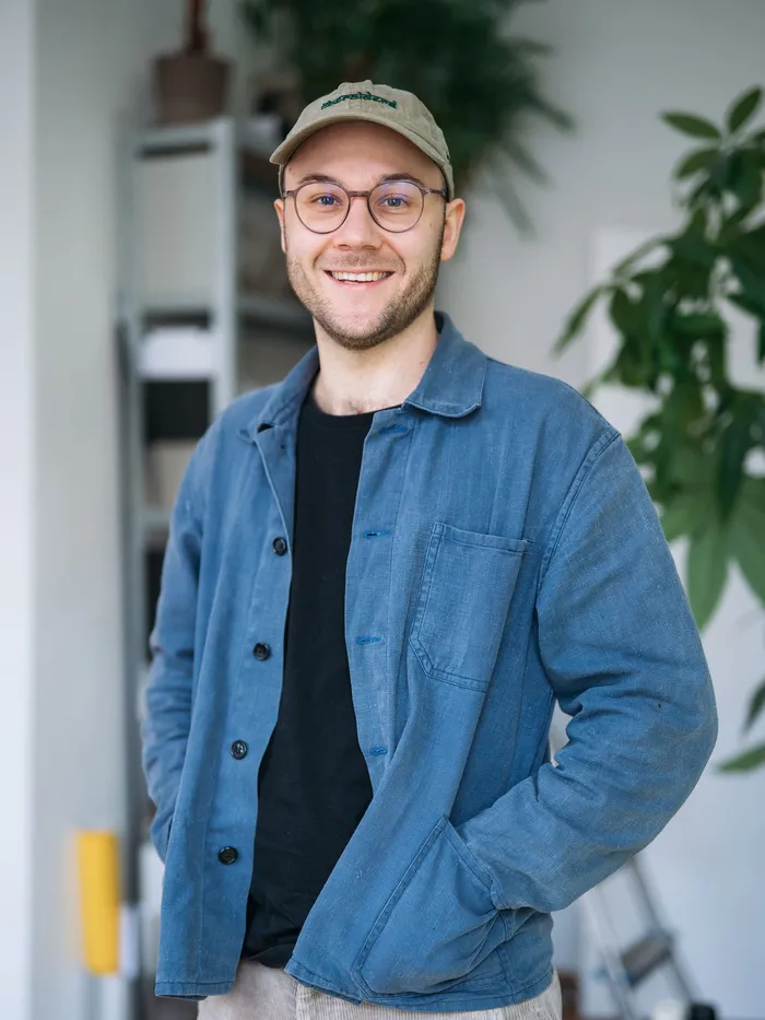 Portrait of a smiling man wearing glasses and a cap, standing indoors with plants in the background.