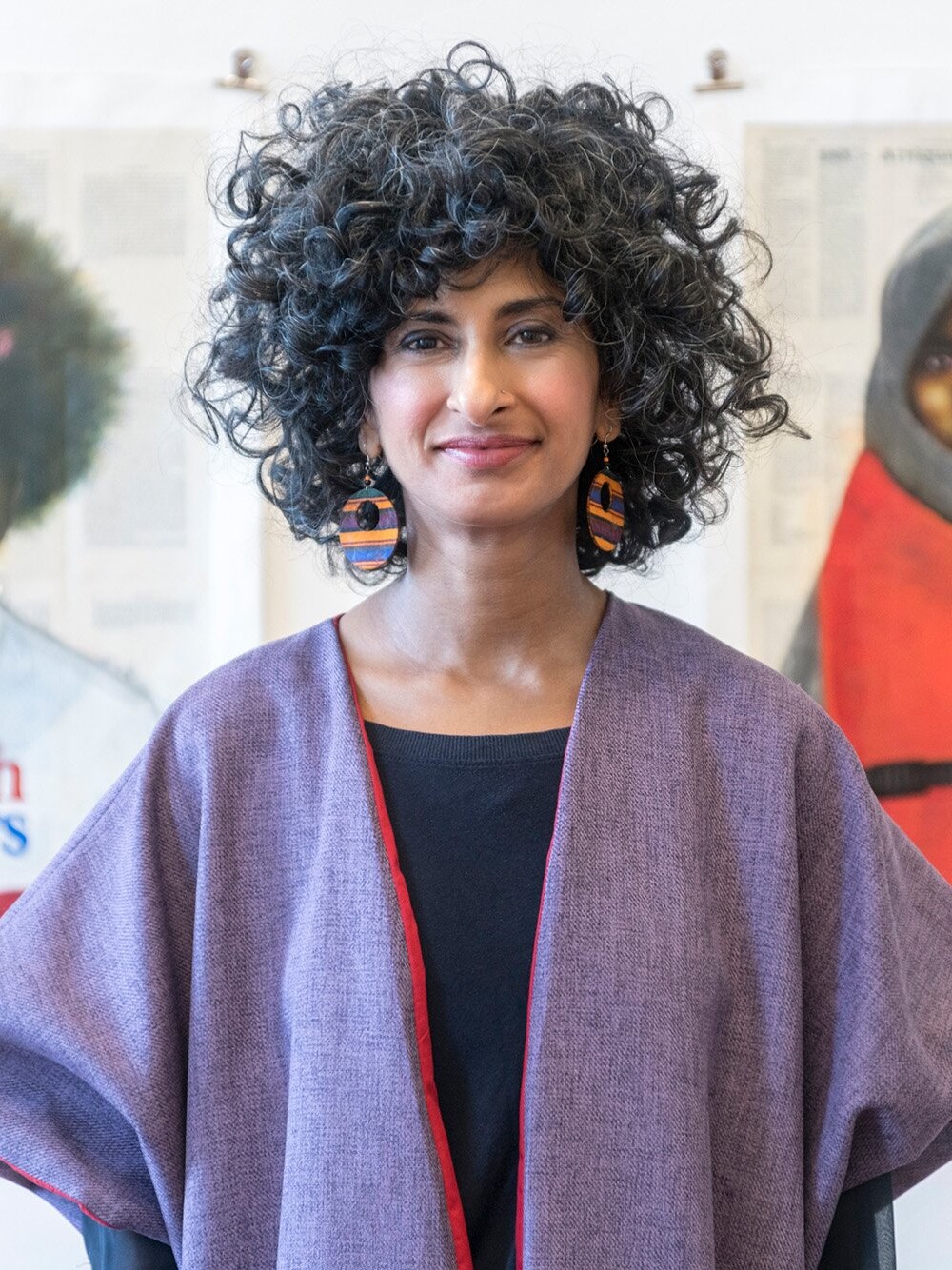 Porträt einer Frau mit lockigem Haar, stehend vor großformatigen Bildern an einer Wand, trägt ein weites Oberteil.