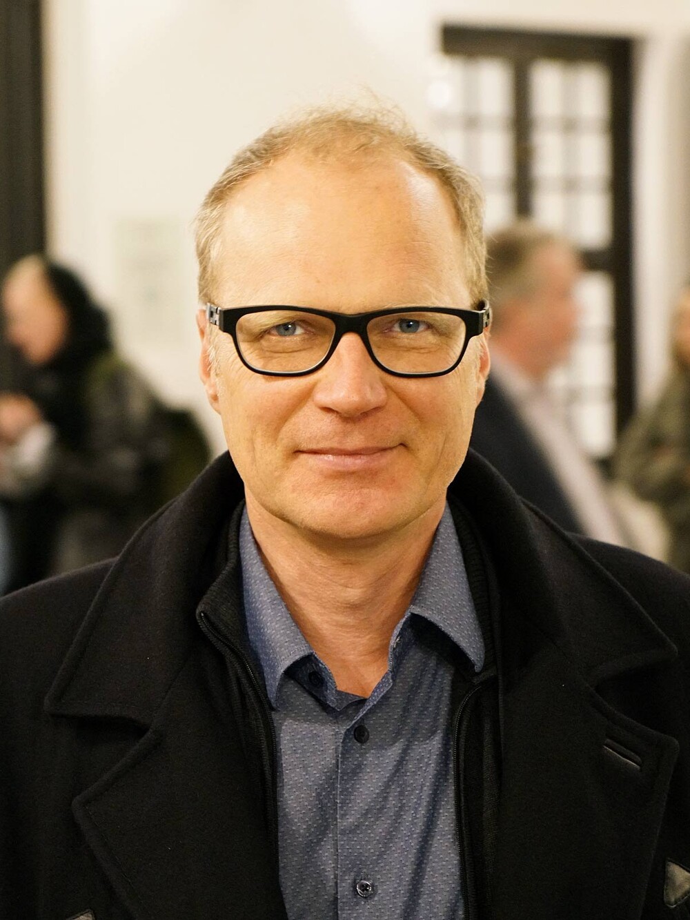 Portrait of a man wearing glasses, standing indoors with several blurred people in the background.