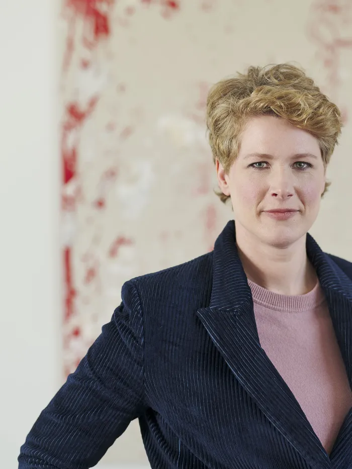 Portrait of a woman with short blonde hair wearing a dark jacket over a pink top, standing in front of a large abstract painting with red and white splashes.