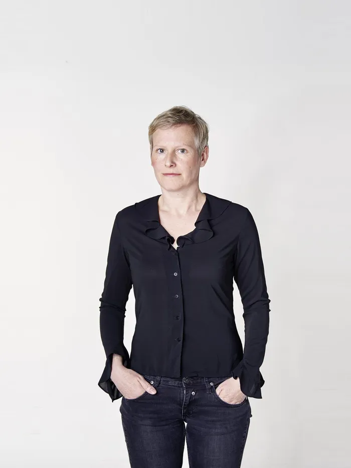 Portrait of a person with short light hair standing against a light background, wearing a dark top and jeans.