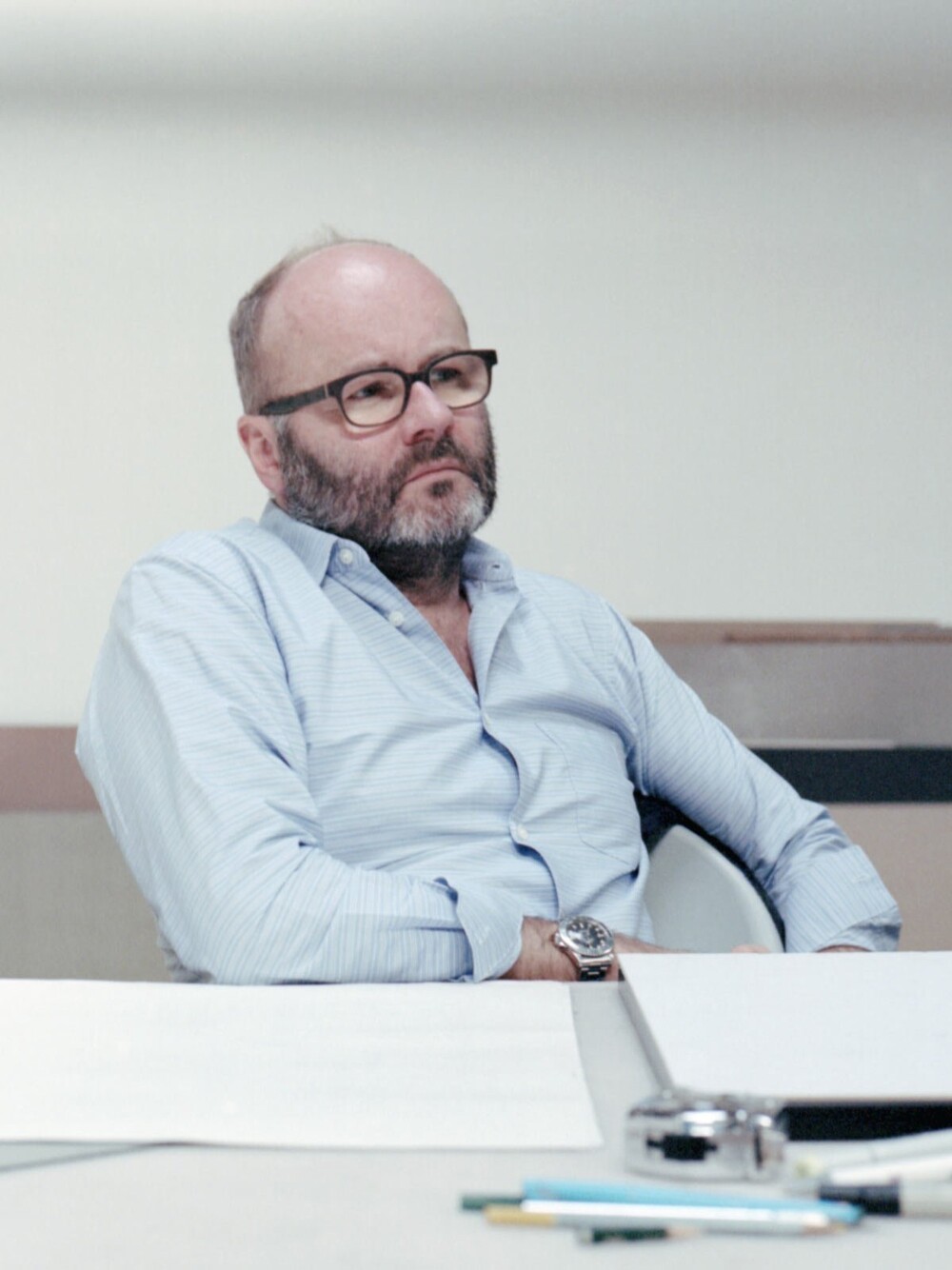 Porträt eines Mannes mit Brille und Bart, sitzend an einem Tisch mit Papieren und Zeichenwerkzeugen in einem Innenraum.