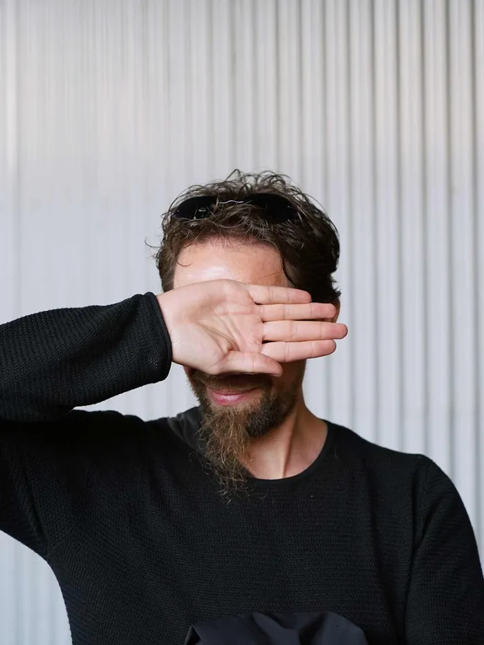 Man with a beard partially covering his face with his hand, standing in front of a light-colored ribbed wall.