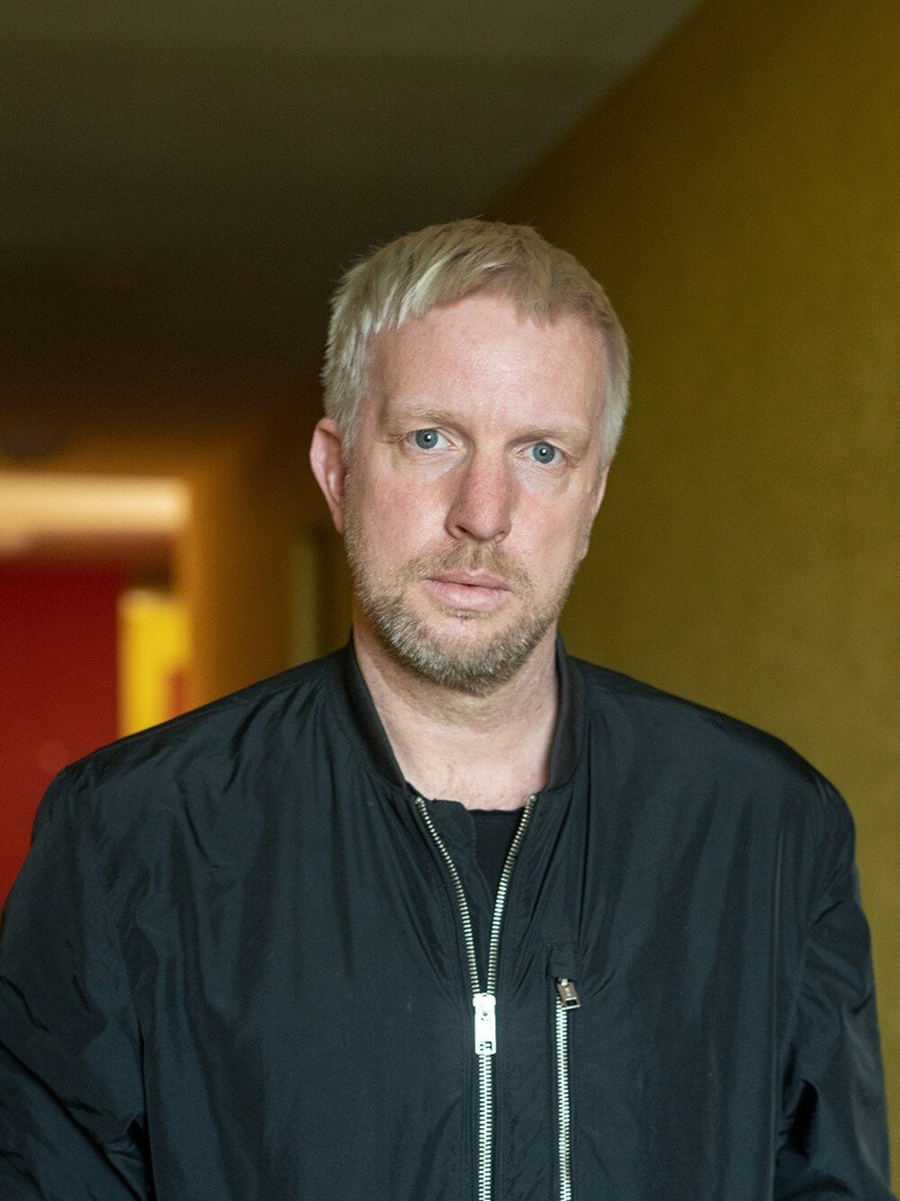 Portrait of a man with short light hair and a beard, wearing a dark jacket and standing indoors.