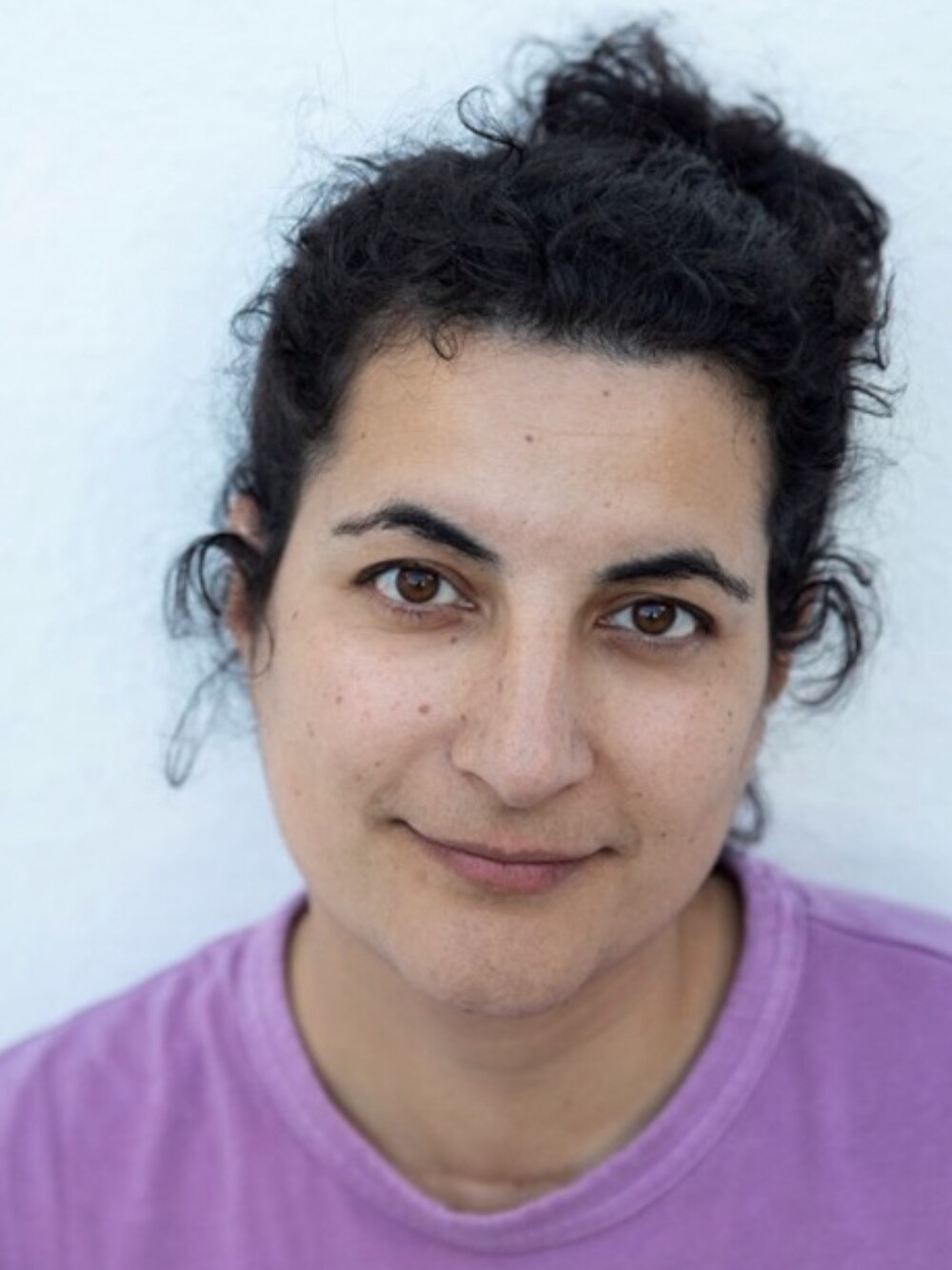 Nahporträt einer Person mit dunklem, lockigem Haar vor hellem Hintergrund, trägt ein hellviolettes Oberteil.