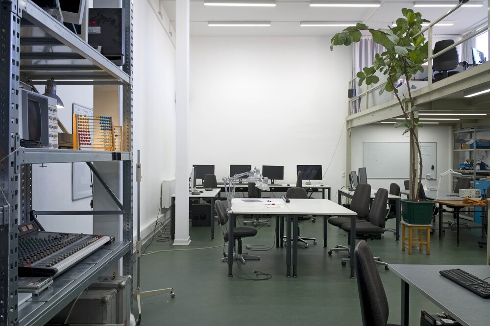 A high-ceilinged room with tables and computers, a shelf on the wall, and a plant under the gallery.