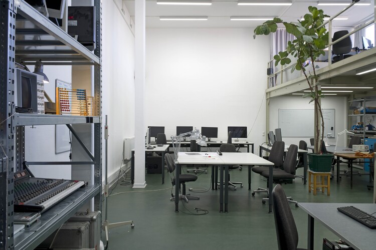 A high-ceilinged room with tables and computers, a shelf on the wall, and a plant under the gallery.