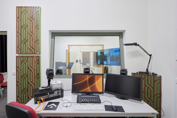 A computer on a desk in front of a window looking into another room.