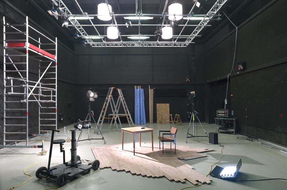 A room with scaffolding, lighting trusses on the ceiling, a table and chair in the middle, and a film dolly next to it.