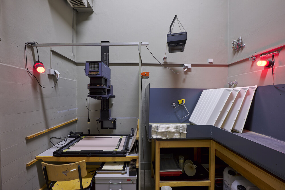 A photography darkroom with an enlarger on a worktable, red safelights mounted on the walls, and work surfaces holding developing trays and equipment.
