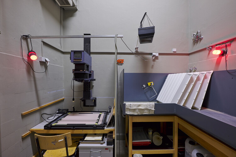 A photography darkroom with an enlarger on a worktable, red safelights mounted on the walls, and work surfaces holding developing trays and equipment.