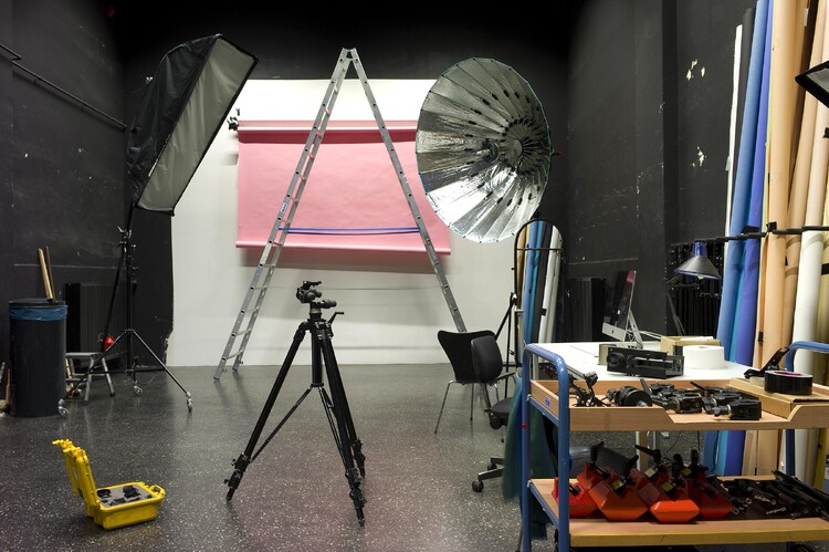 A pink roll of paper hangs on a white wall, in front of it a ladder, a tripod, and an open camera bag.