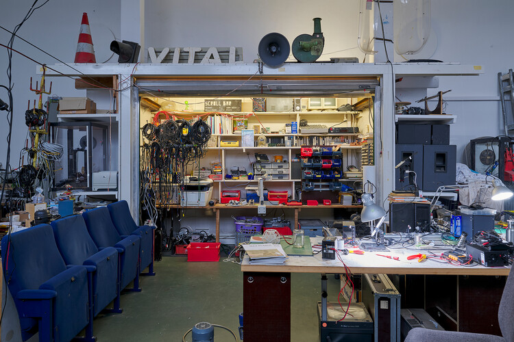 A fully equipped workshop with a large workbench in the foreground holding tools, cables, and electronic components; shelves with equipment, spare parts, and coiled wires in the background, and rows of seats along one side.