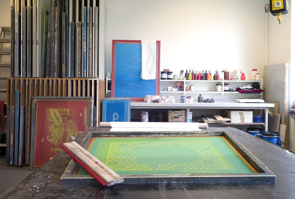 A coated screen with a squeegee in the foreground, with tubes of paint and additional screens behind it.