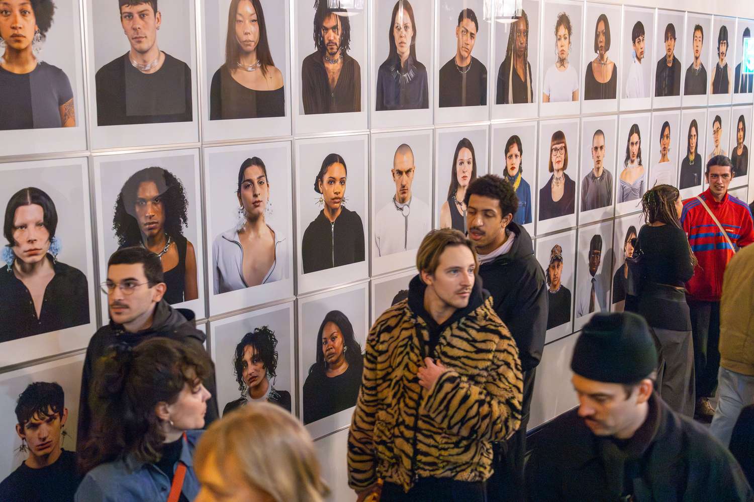 Many people stand in front of a wall with portrait photographs