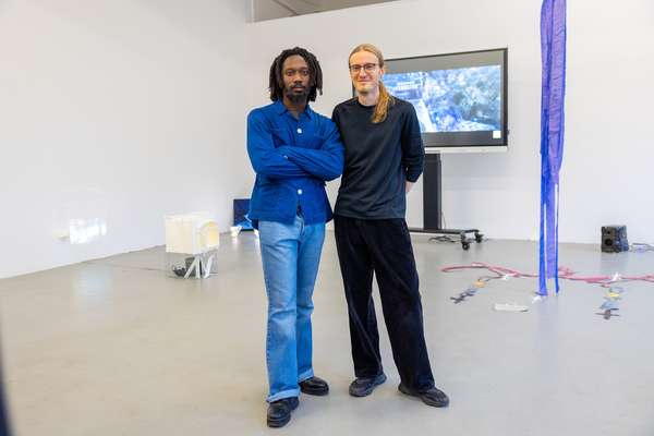 Zwei Männer stehen in einem Ausstellungsraum mit mehren Objekten und einem großen Bildschirm im Hintergrund nebeneinander. Der linke Mann ist dunkelhäutig, trägt einen Bart und ein blaues Hemd sowie eine Jeans. Der rechte Mann trägt seine blonden langen Haare zu einem Zopf gebunden. Er trägt eine Brille und schwarze Kleidung. Er lächelt.